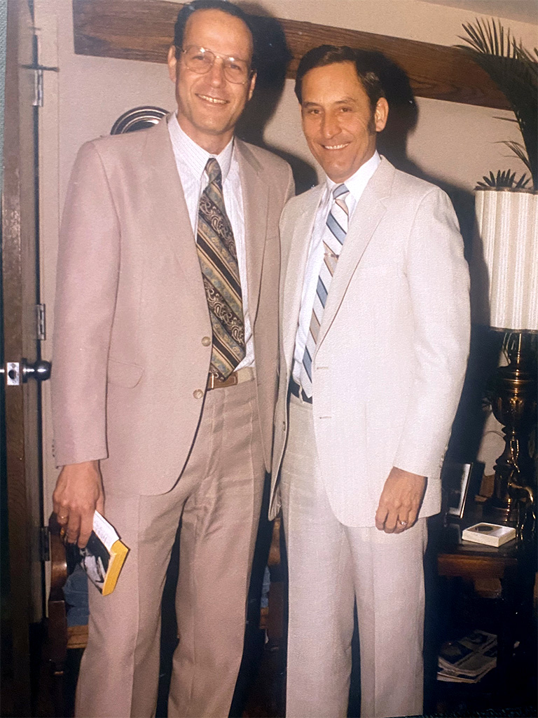 Two men in casual suits stand close together smiling and posing for the camera.